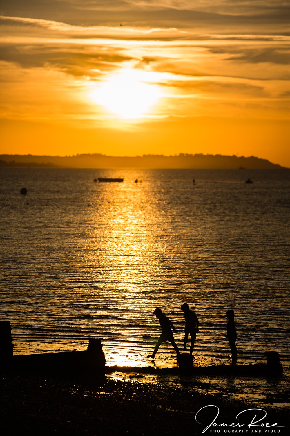 Silhouettes of three children walking along a beach at sunset, with the sun reflecting on the water, boats in the distance, and a cloudy sky.
