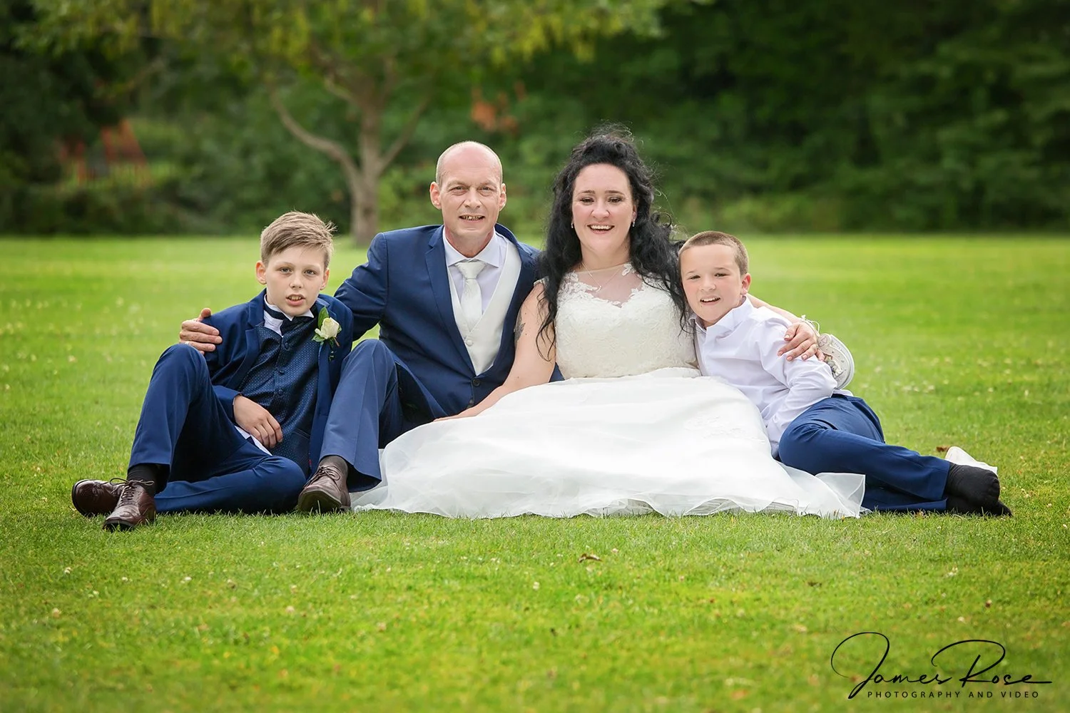 A family of four sitting on grass in a park, dressed in formal wedding attire, smiling at the camera. The woman is in a white wedding dress, and the man and boys are in suits.