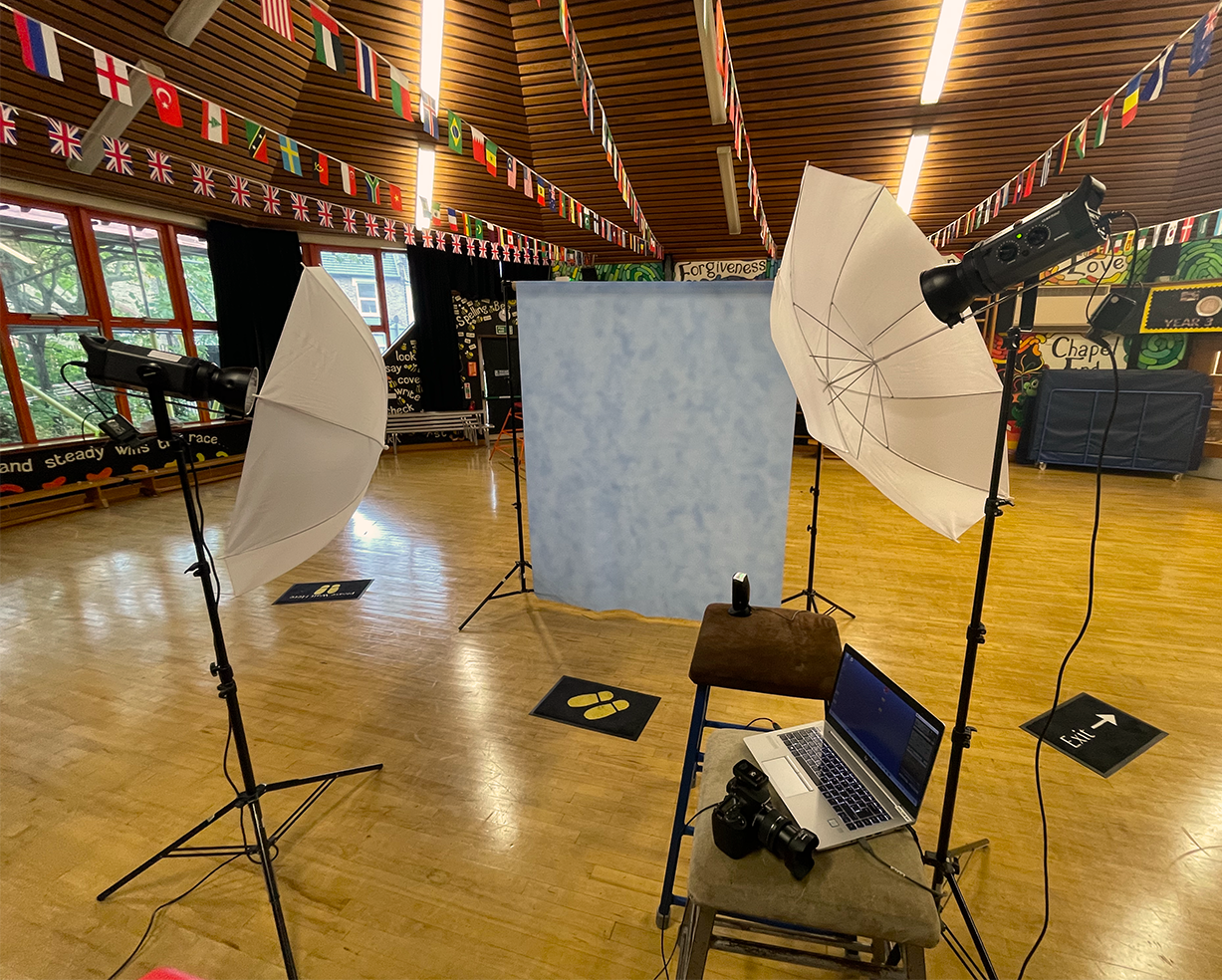 Photography studio setup with two umbrella lights, a laptop with a camera, CCTV, and a backdrop in a room decorated with international flags hanging from the ceiling.