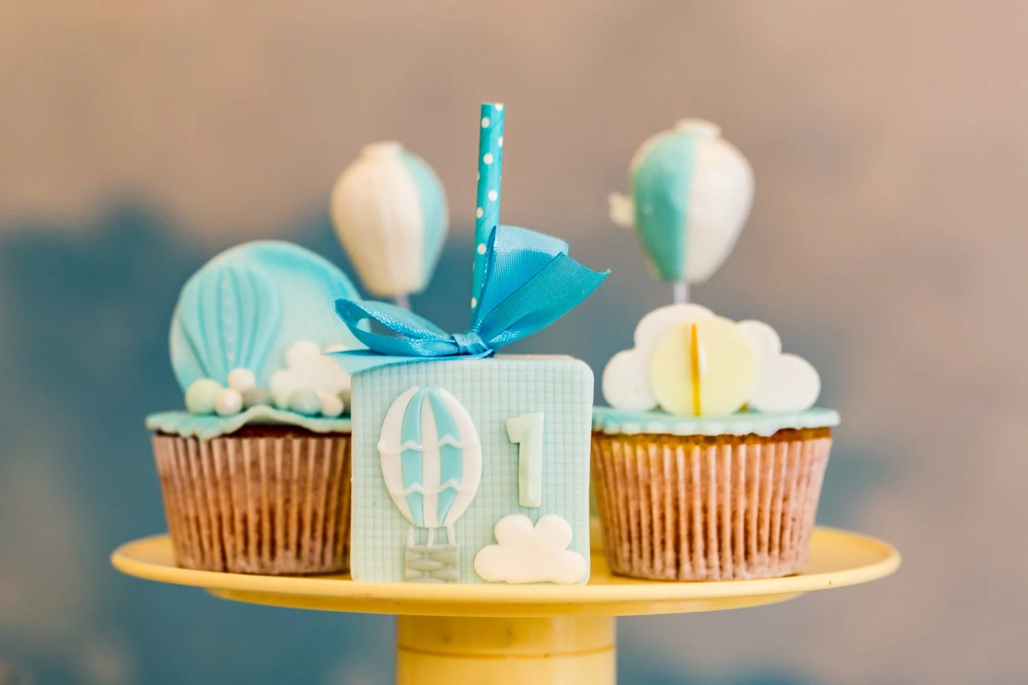 Cupcakes decorated with pastel blue, yellow, and white themes featuring balloons, clouds, and a number one, suited for a first birthday celebration.