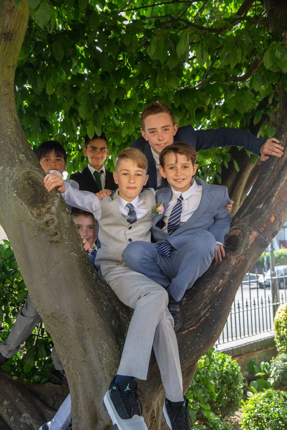 Group of children in formal attire sitting and standing on a large tree branch, smiling and posing for the photo.