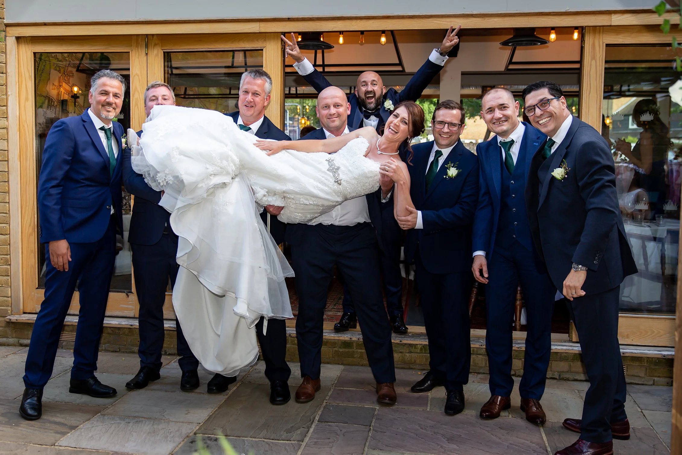Group of men in suits holding a bride in a white wedding dress outside a building with wooden doors and large windows.