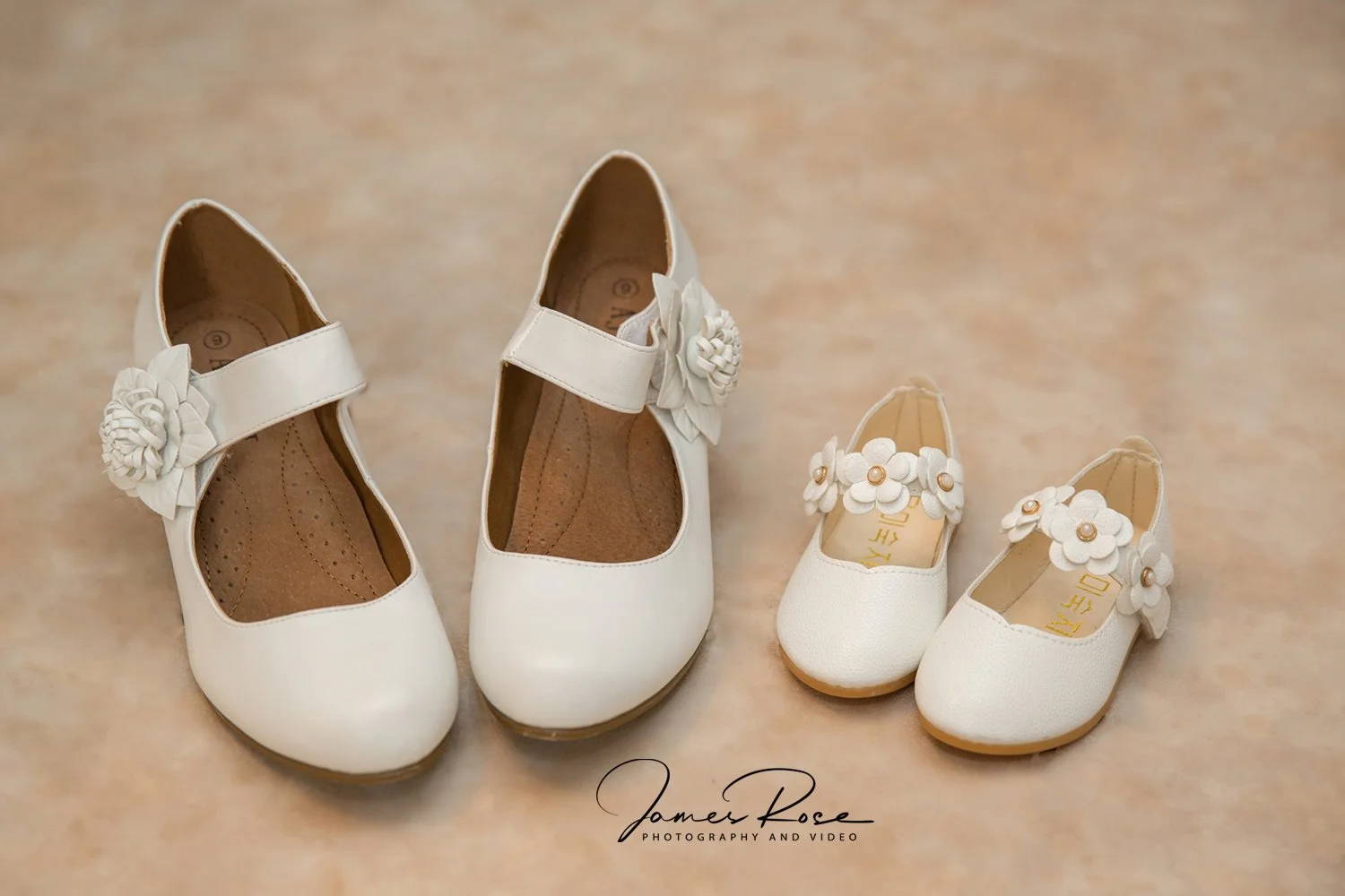 Pair of white girl's shoes with floral embellishments, one larger pair on the left and a smaller pair on the right, placed on a beige surface.