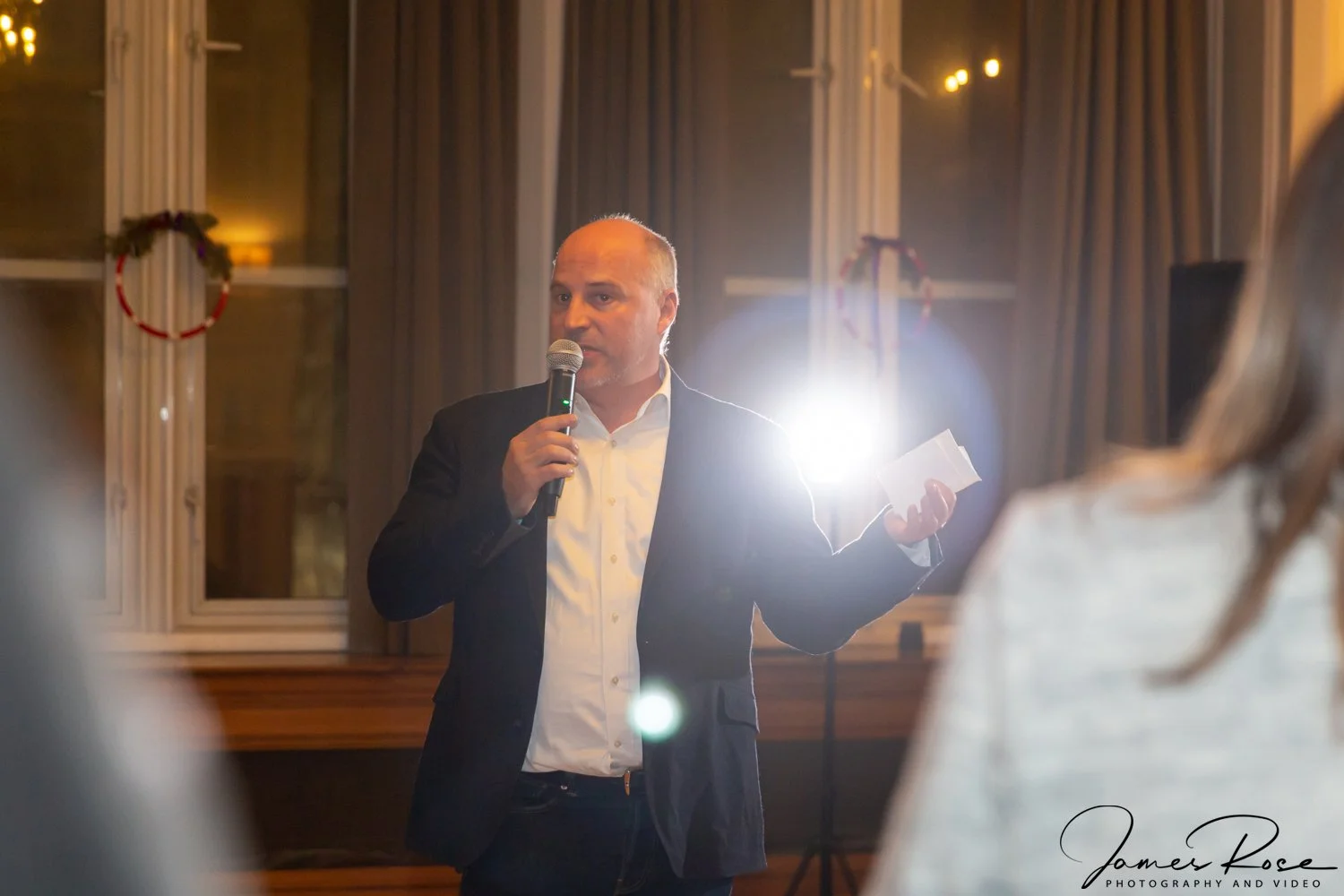 A man in a suit holding a microphone and a piece of paper, speaking at an indoor event with festive decorations, walls with large windows, and warm lighting.