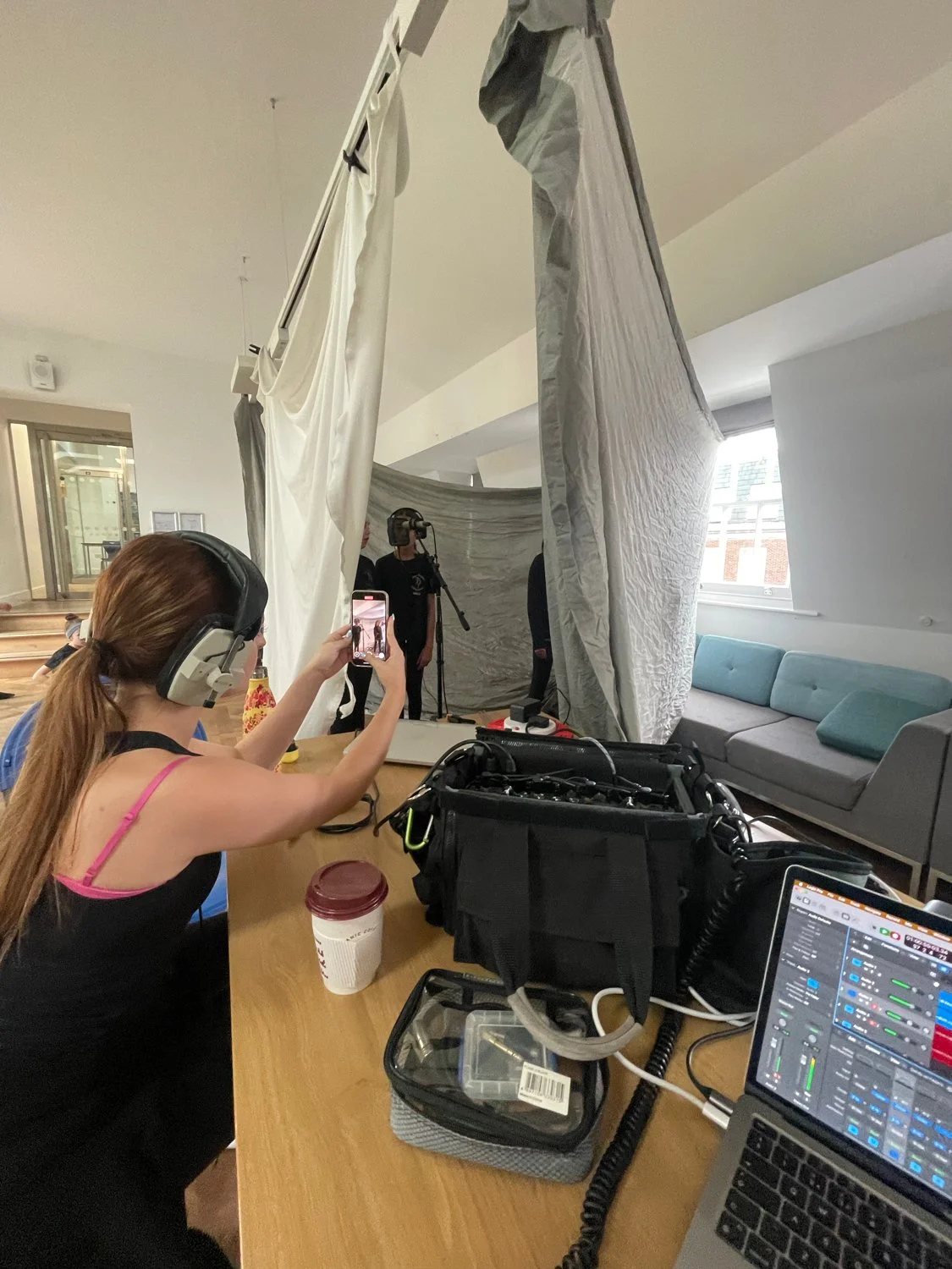 A woman with headphones is taking a photo with her phone of a filming setup with cameras and curtains, and a person is in the background.