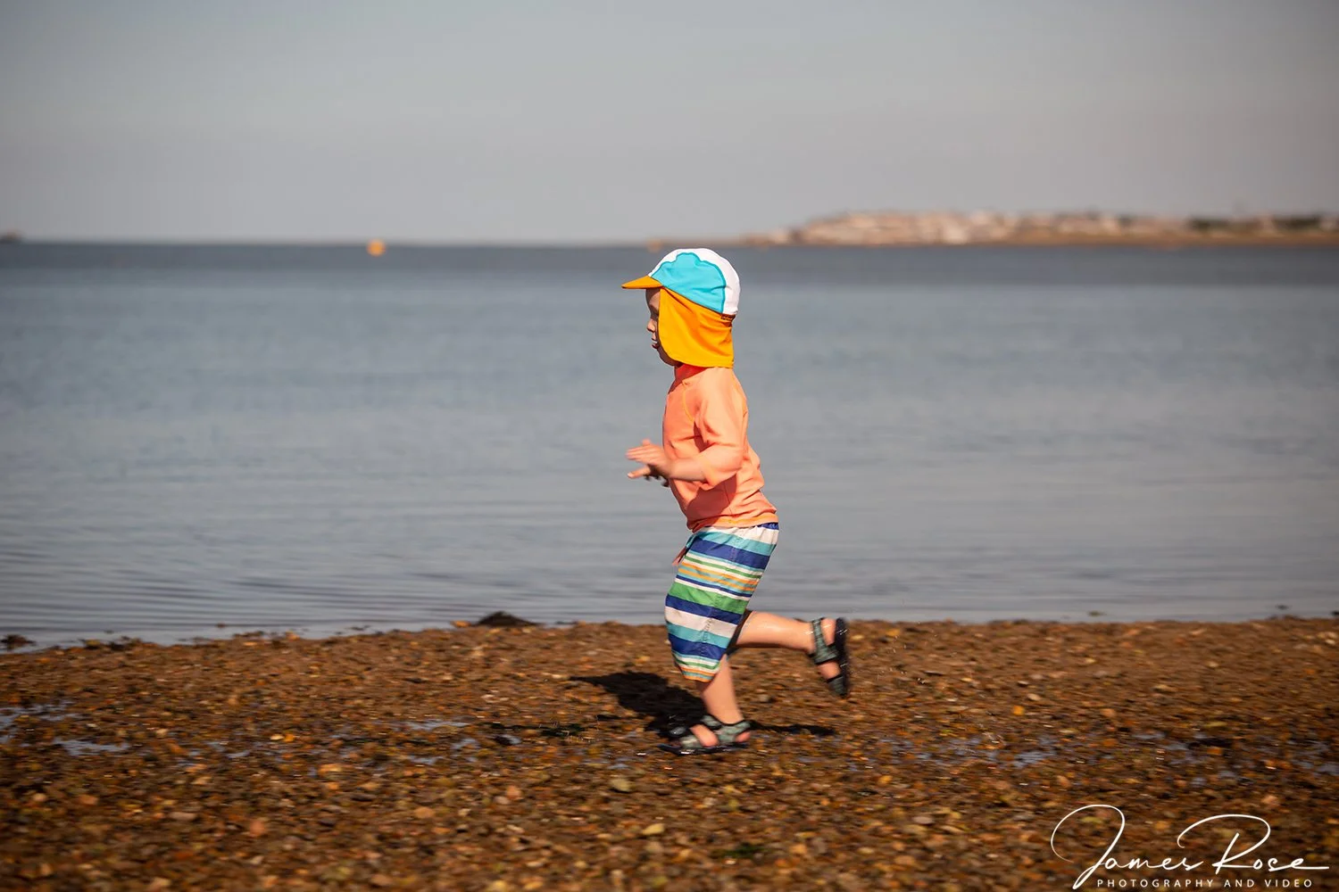 A young boy running along the shoreline on a rocky beach, wearing a colorful hooded hat, an orange shirt, and striped shorts, with water and distant land in the background.