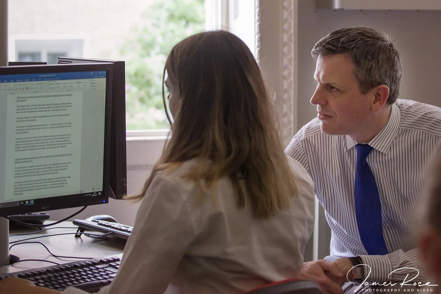 A man in a striped shirt and blue tie looking at a woman with long hair sitting at a computer workstation in an office.