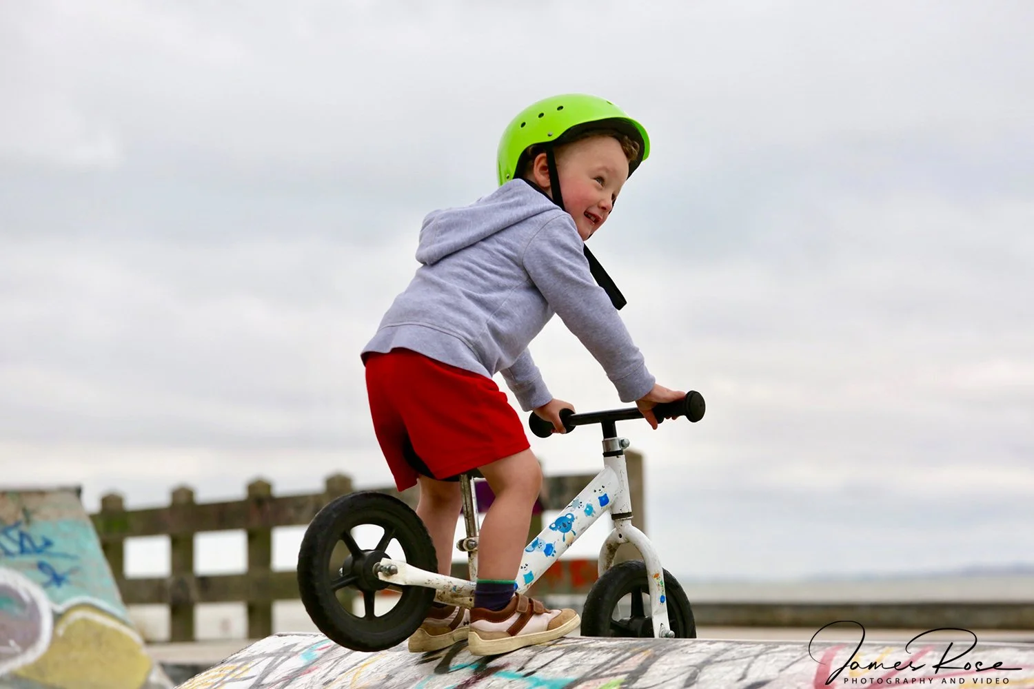 A young boy wearing a green helmet, gray hoodie, red shorts, and sandals riding a white balance bike at a skate park with graffiti-covered ramps.