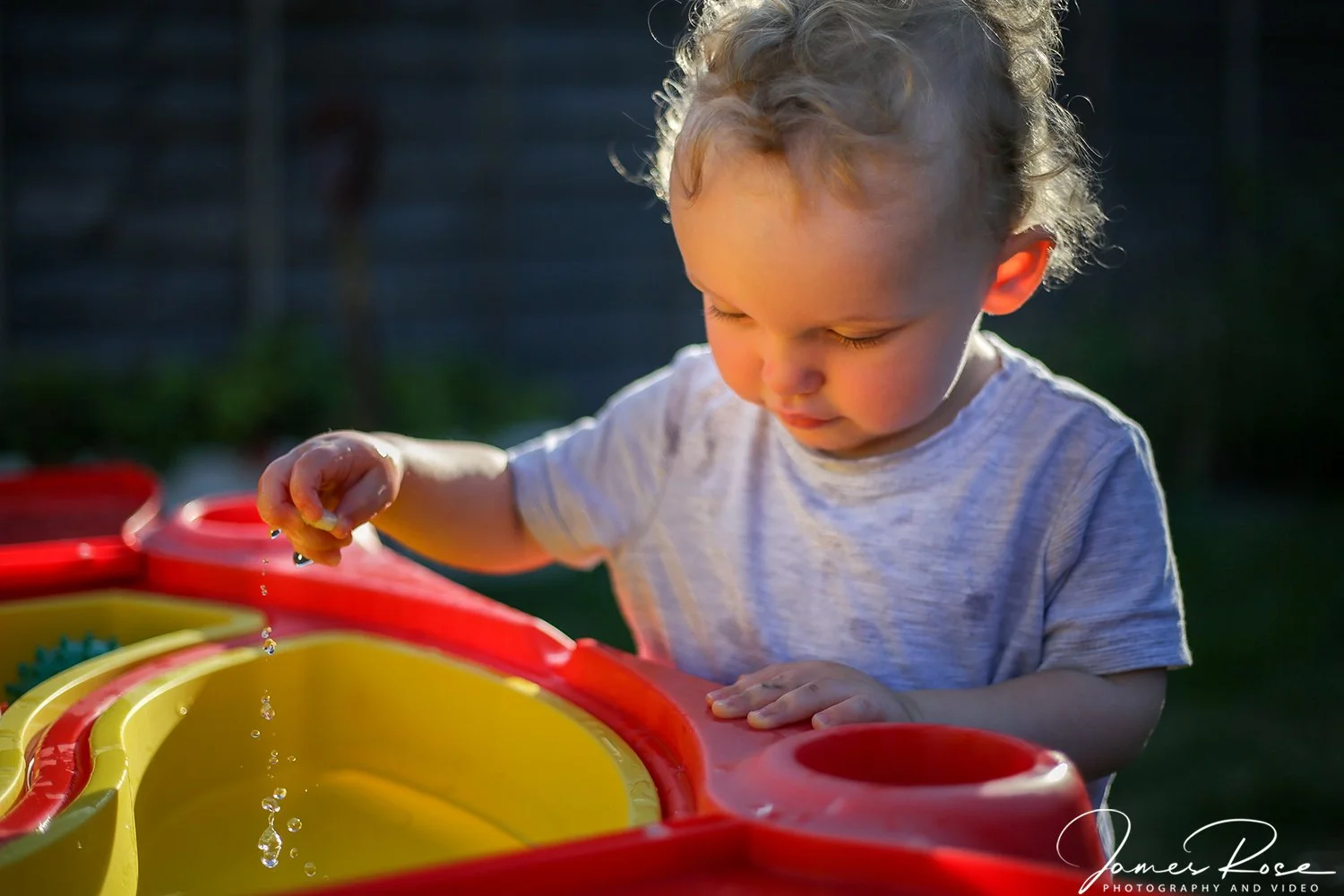 A young boy with curly blond hair playing with water in a red and yellow water table outdoors during the daytime.