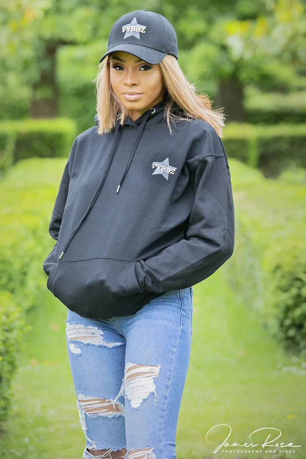 A young woman with blonde hair wearing a black hoodie and matching black baseball cap with a logo, standing outdoors in a green park.