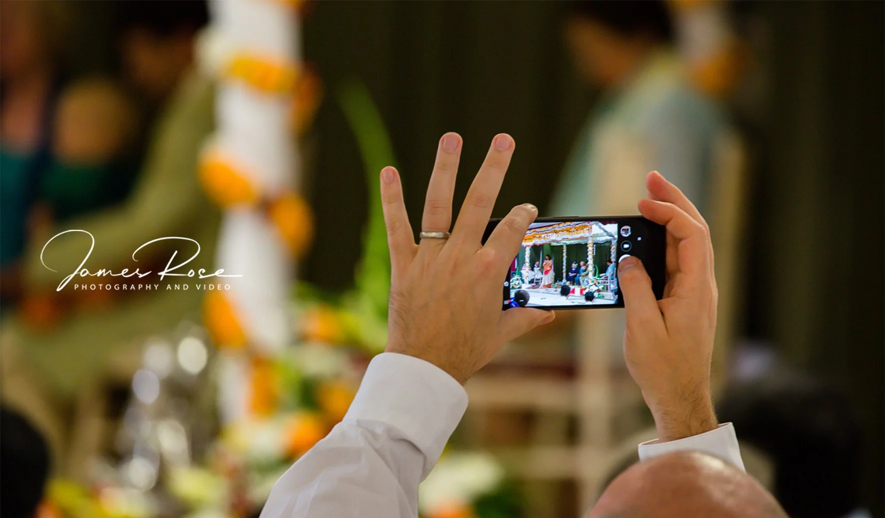 Person holding a smartphone taking a picture of a stage with performers at a celebration or ceremony, decorated with flowers and colorful elements.