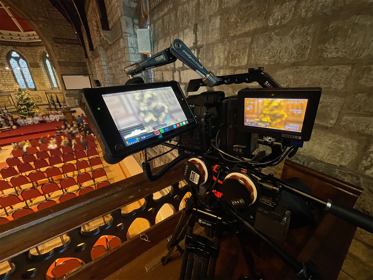 A professional video camera setup on a tripod, recording inside a church or auditorium with red chairs, a Christmas tree, and stained glass windows.
