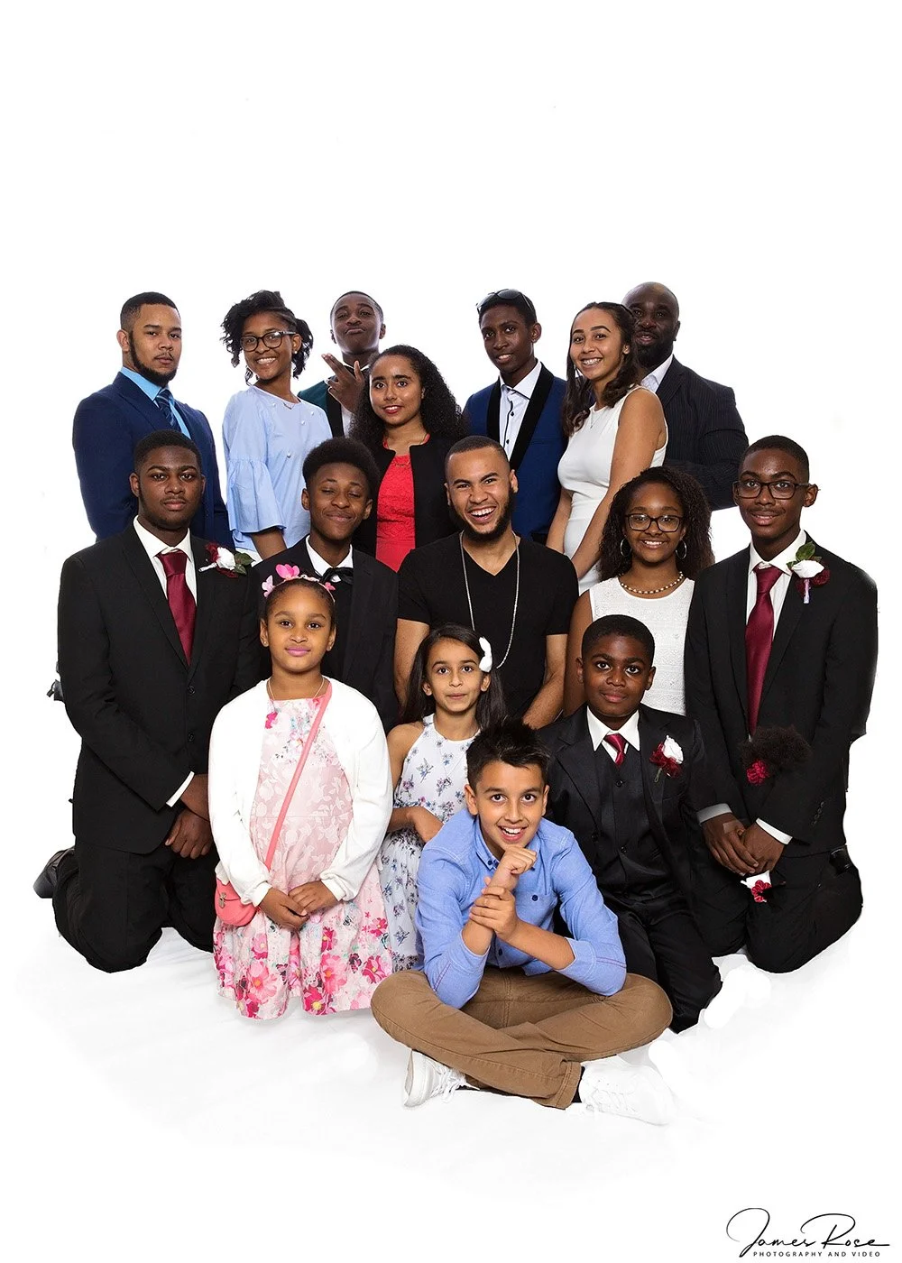 A large group of diverse people, including children, teenagers, and adults, dressed in formal attire, gathered for a group photo against a white background, smiling and posing happily.