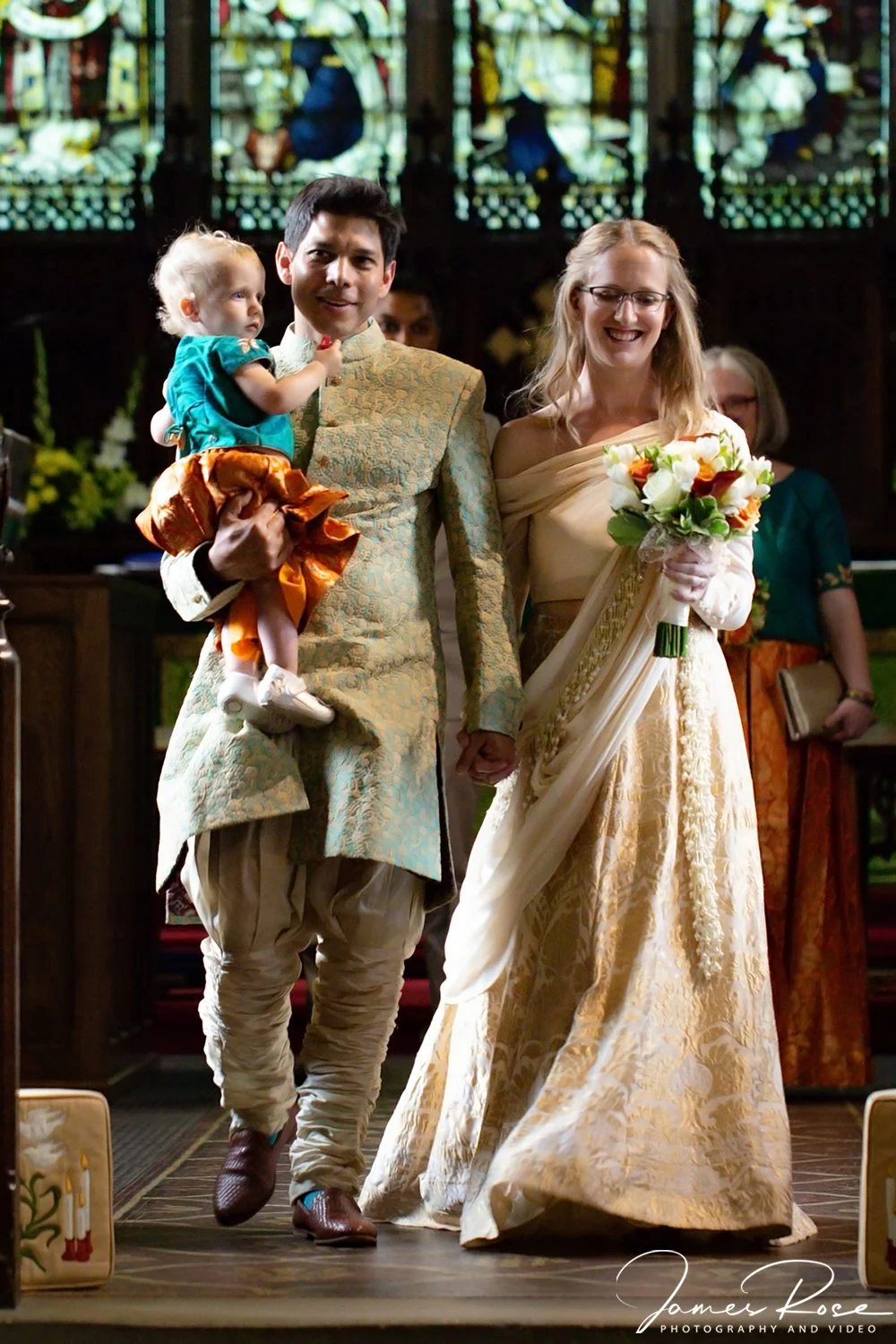 A couple dressed in traditional or wedding attire holding hands, with the woman holding a bouquet of flowers, walking down an aisle in a church with stained glass windows, accompanied by a small child.