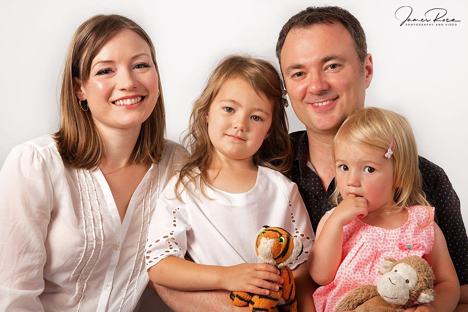 A family of four smiling for a portrait against a white background. The mother has shoulder-length brown hair and wears a white blouse, the father has short brown hair and wears a black shirt. Their two daughters have blonde hair; one girl is holding