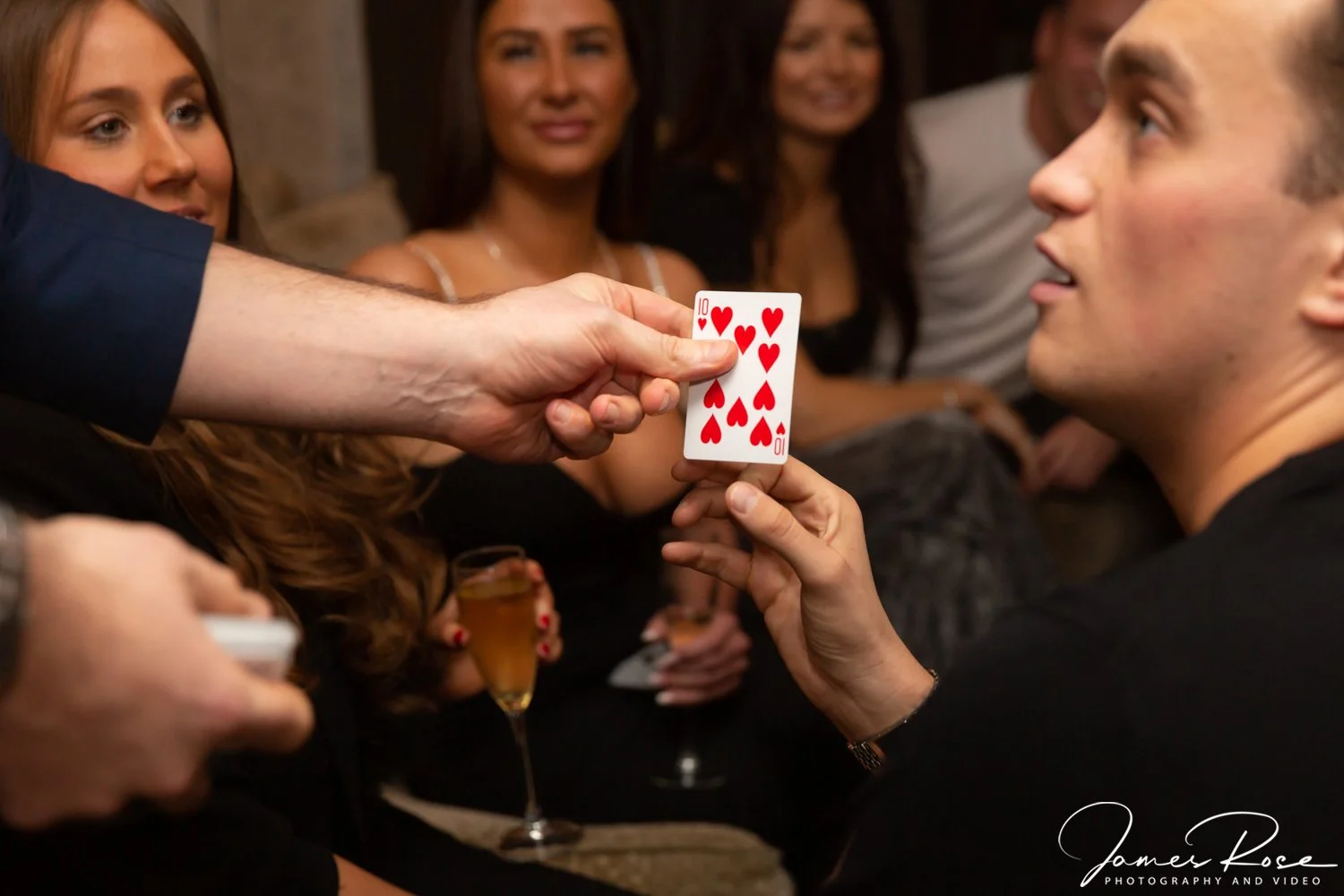 A group of people at a party, with one person holding a winning ten of hearts playing card, as another person points to it. The group appears to be celebrating or enjoying themselves.