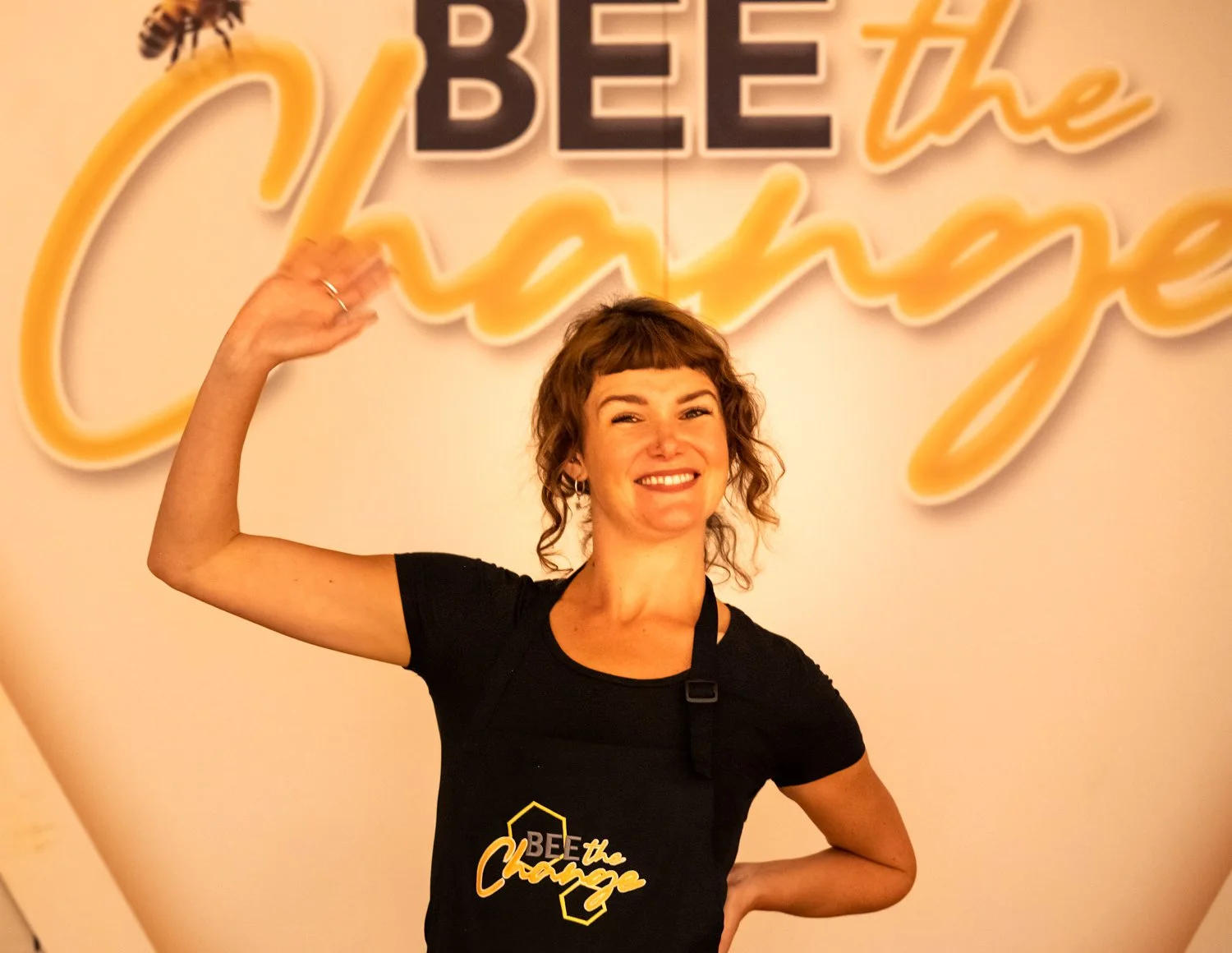 A woman with curly brown hair smiling and flexing her right arm, standing in front of a sign with the words 'BE the Change' and a bee illustration, wearing a black shirt with the same phrase on it.