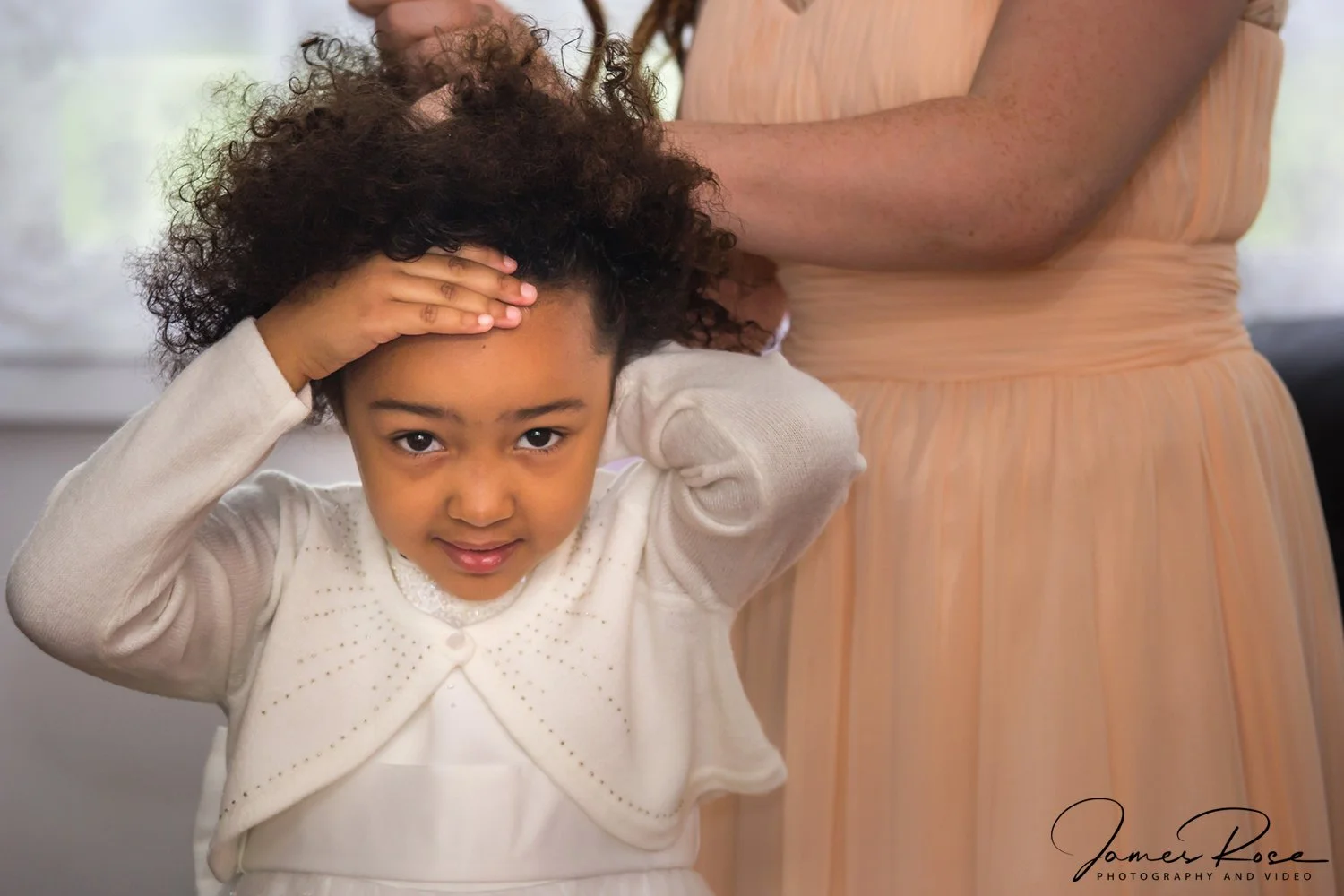 A young girl with curly hair adjusting her hat while standing beside an adult woman in a peach-colored dress.