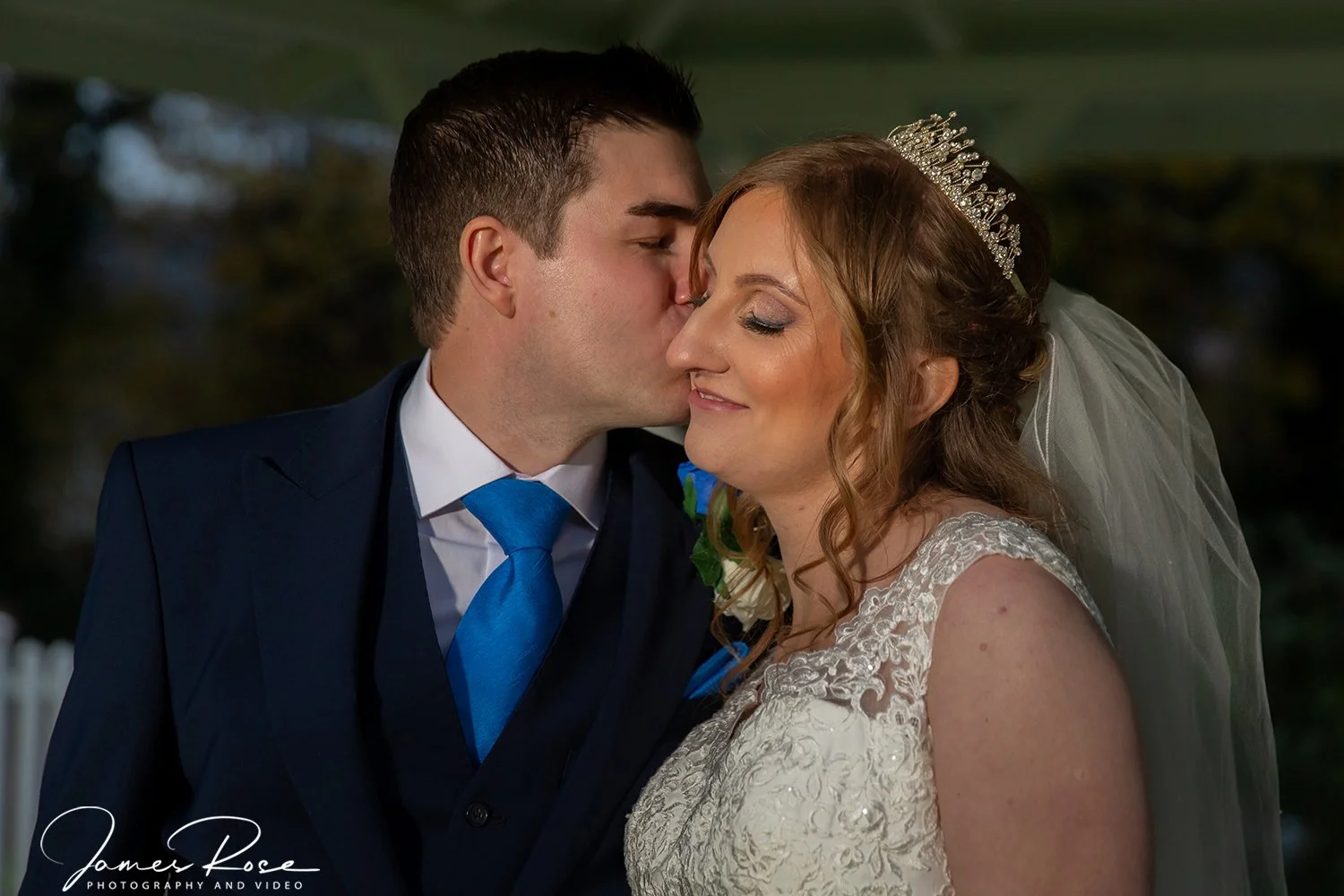 A groom kissing a bride on the forehead during their wedding ceremony, with the bride wearing a lace wedding dress and tiara, and the groom in a navy suit with a bright blue tie.