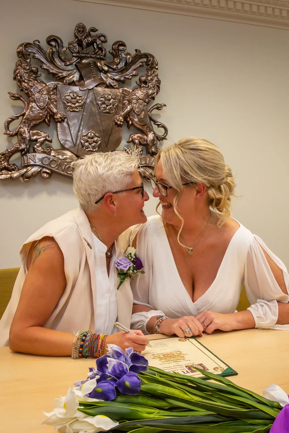 Two women, one with short white hair and the other with blonde hair, smiling and leaning close together at a table with a wedding certificate and purple flowers, in front of a coat of arms on the wall.