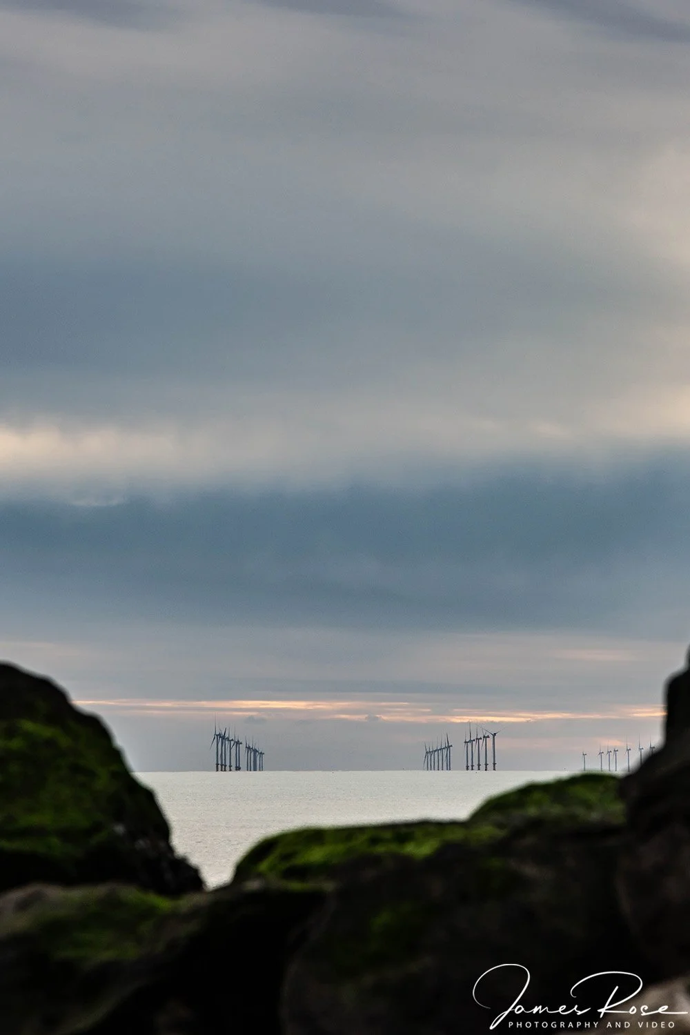 Overcast sky over the ocean with wind turbines in the distance, partially obscured by moss-covered rocks in the foreground.