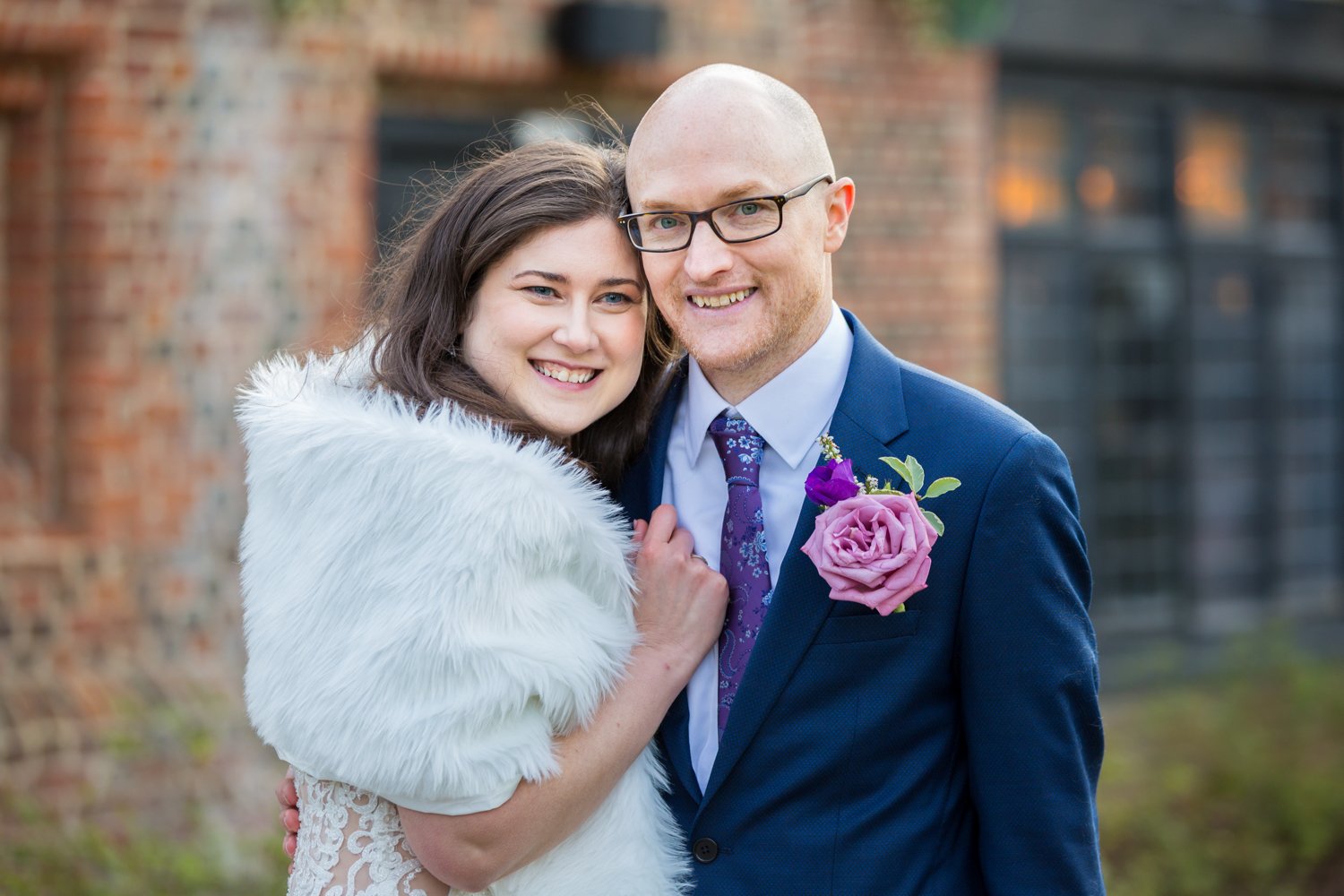 A happy couple in wedding attire, outside in front of a brick building. The woman is wearing a white fur shawl and lace dress, and the man is in a blue suit with a pink flower boutonniere and a purple tie, both smiling.