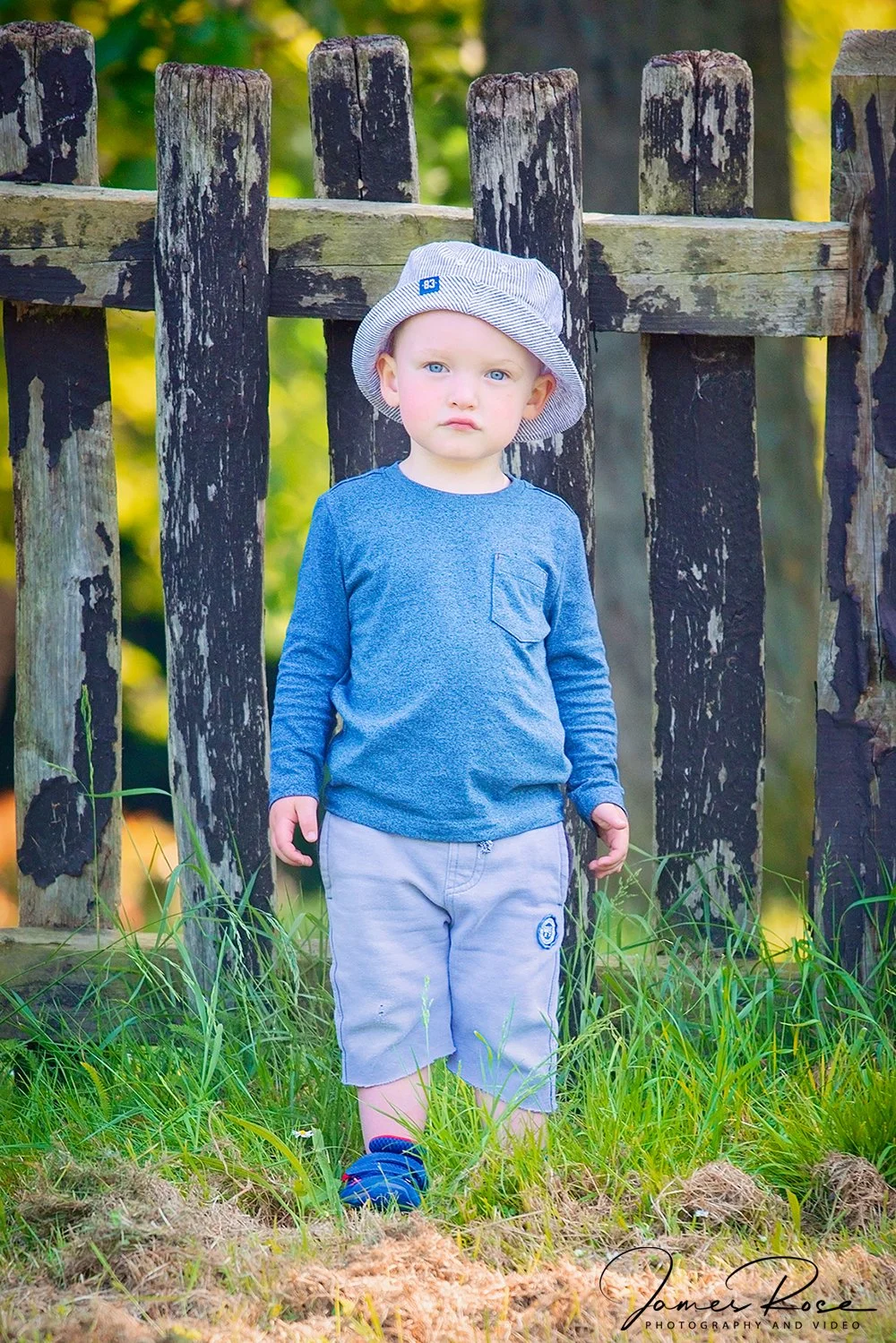 A young boy with blonde hair and blue eyes stands outdoors in front of a weathered wooden fence, wearing a straw hat, a blue long-sleeve shirt, and gray shorts, with green grass at his feet.