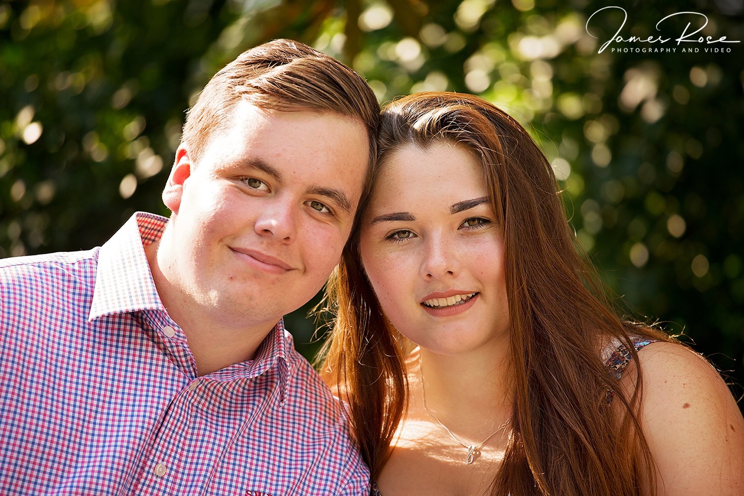 A young man and woman smiling outdoors in a sunny setting with green foliage in the background.