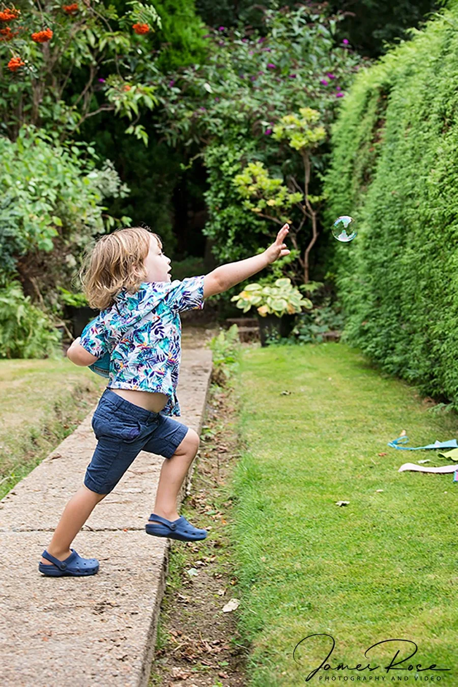A young boy with blonde hair, wearing a colorful short-sleeved shirt, blue shorts, and blue Crocs, reaching out to touch a floating soap bubble in a garden with lush green bushes and plants.