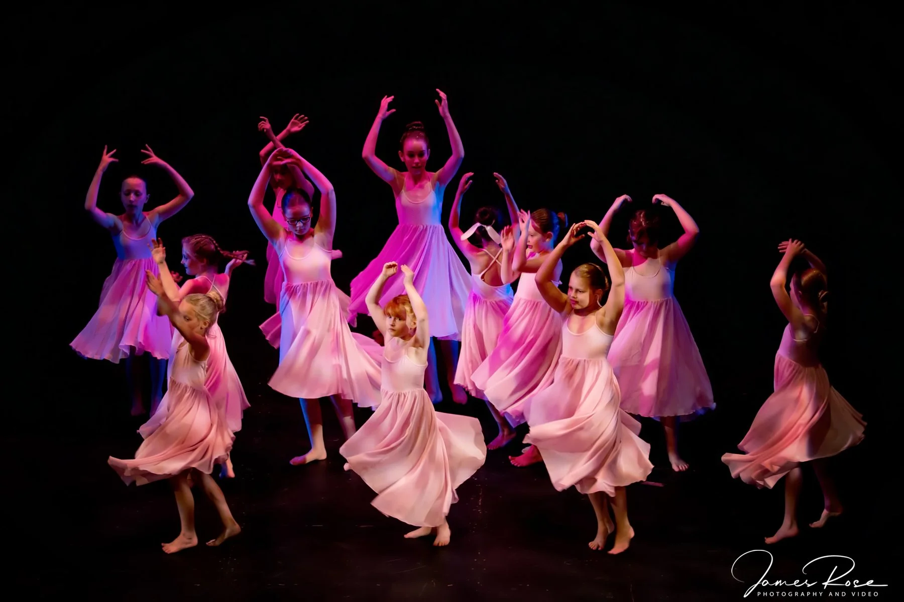 A group of young ballet dancers in pink dresses performing on stage with a dark background.