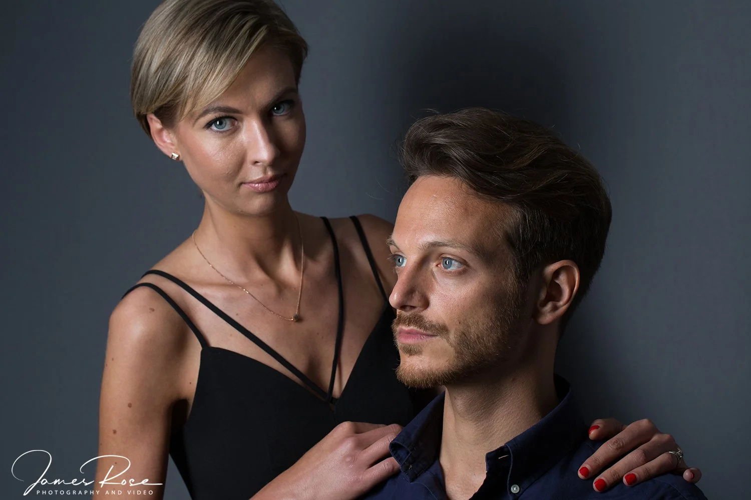 A woman with short blonde hair, wearing a black spaghetti strap dress and small earrings, stands behind a man with dark hair, blue eyes, and a beard, who is seated and wearing a dark shirt. The woman has her hand on the man's shoulder, and they are p