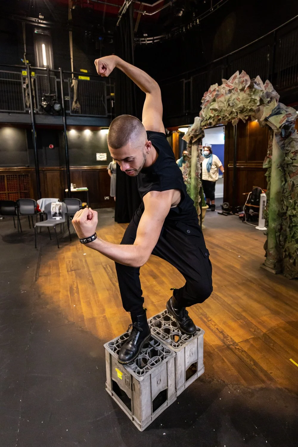Man with a shaved head and tattoos flexing his right arm while balancing on a plastic crate in a room with wooden floors and dark walls, with a decorative arch made of crumpled paper or fabric behind him.