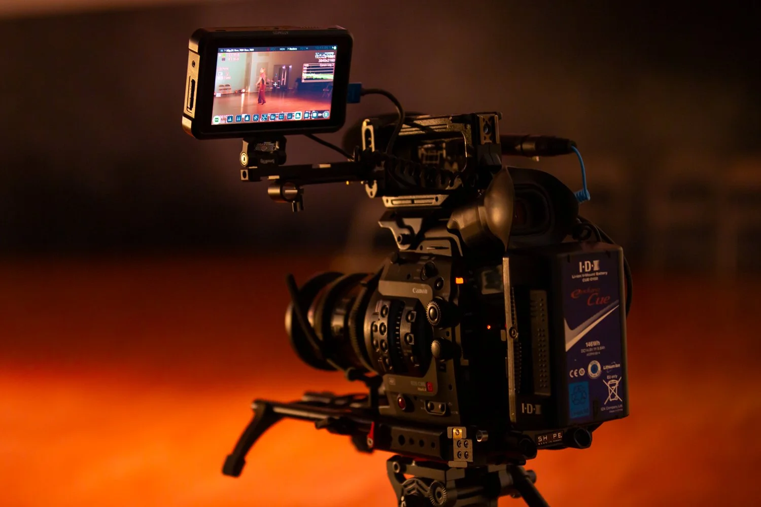 Professional video camera on a tripod with an external monitor, set up in a dimly lit room.