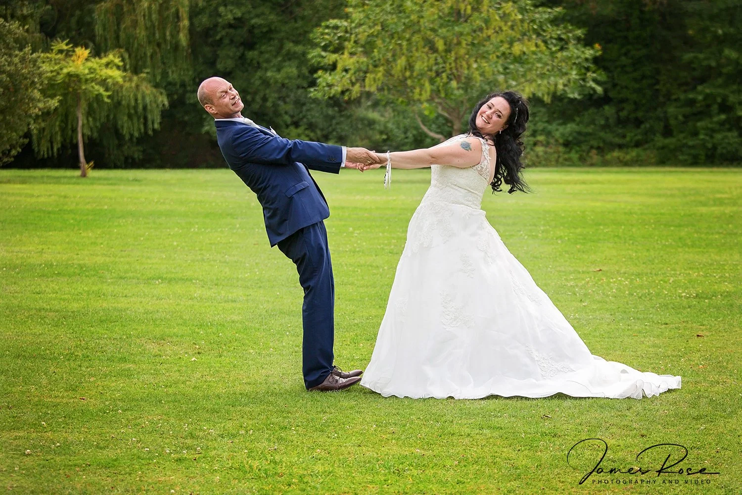 A bride and groom playfully pulling each other in a grassy outdoor setting with green trees in the background. The bride is wearing a wedding dress, and the groom is in a blue suit.