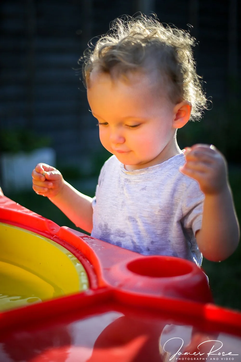 A young boy with curly hair standing outdoors in sunlight, looking at a water table or toy with a focused expression.