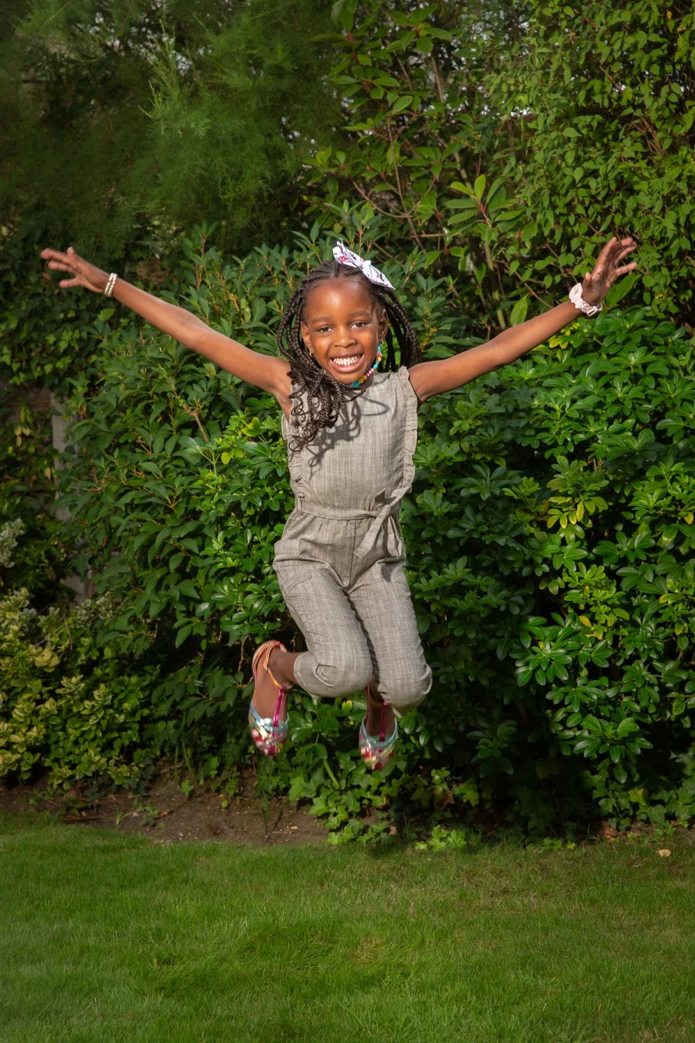 A young girl with braids, wearing a gray sleeveless jumpsuit, is jumping in the air with arms outstretched in a garden with green bushes and trees.