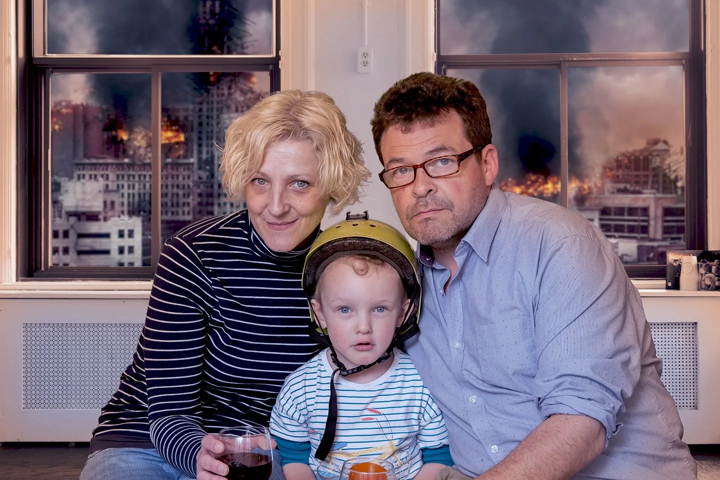 A family of three, including a woman, man, and young boy with a helmet, sitting indoors with a cityscape on fire seen through the windows behind them.