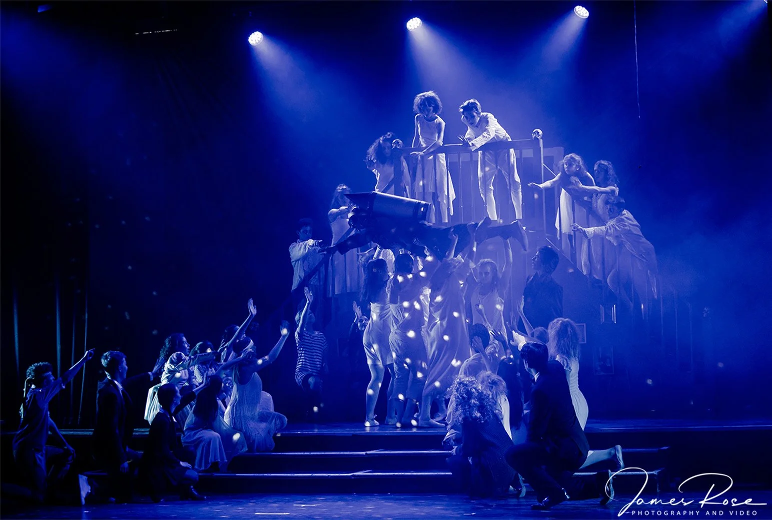 A theatrical stage scene with children and adults, some holding a large prop, bathed in blue lighting, with a multi-level staircase and a railing, suggestive of a play or musical performance.