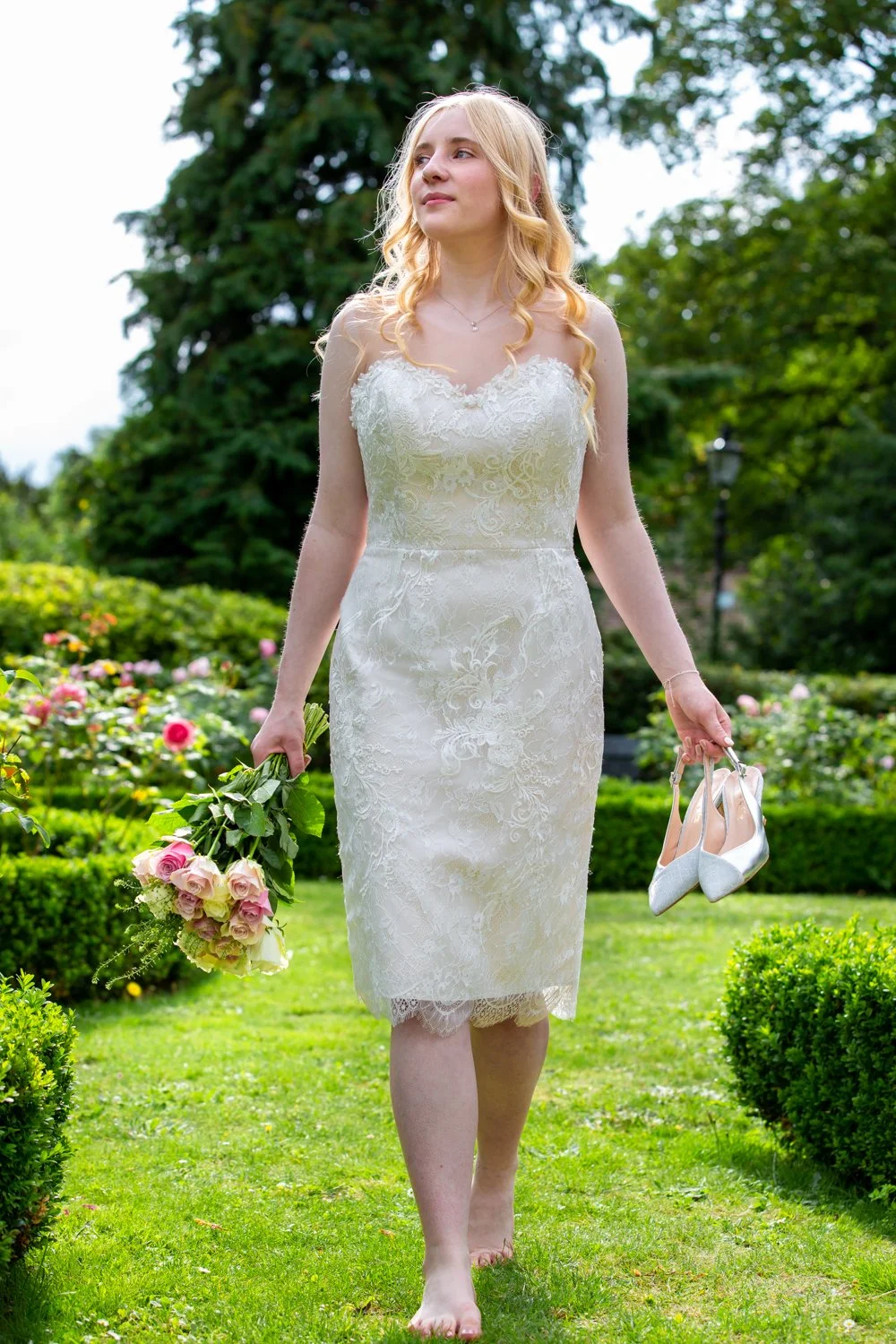 A young woman in a white lace dress holding a bouquet of pink and white roses in one hand and white high-heeled shoes in the other, walking barefoot on a grassy garden path surrounded by green trees and shrubs.