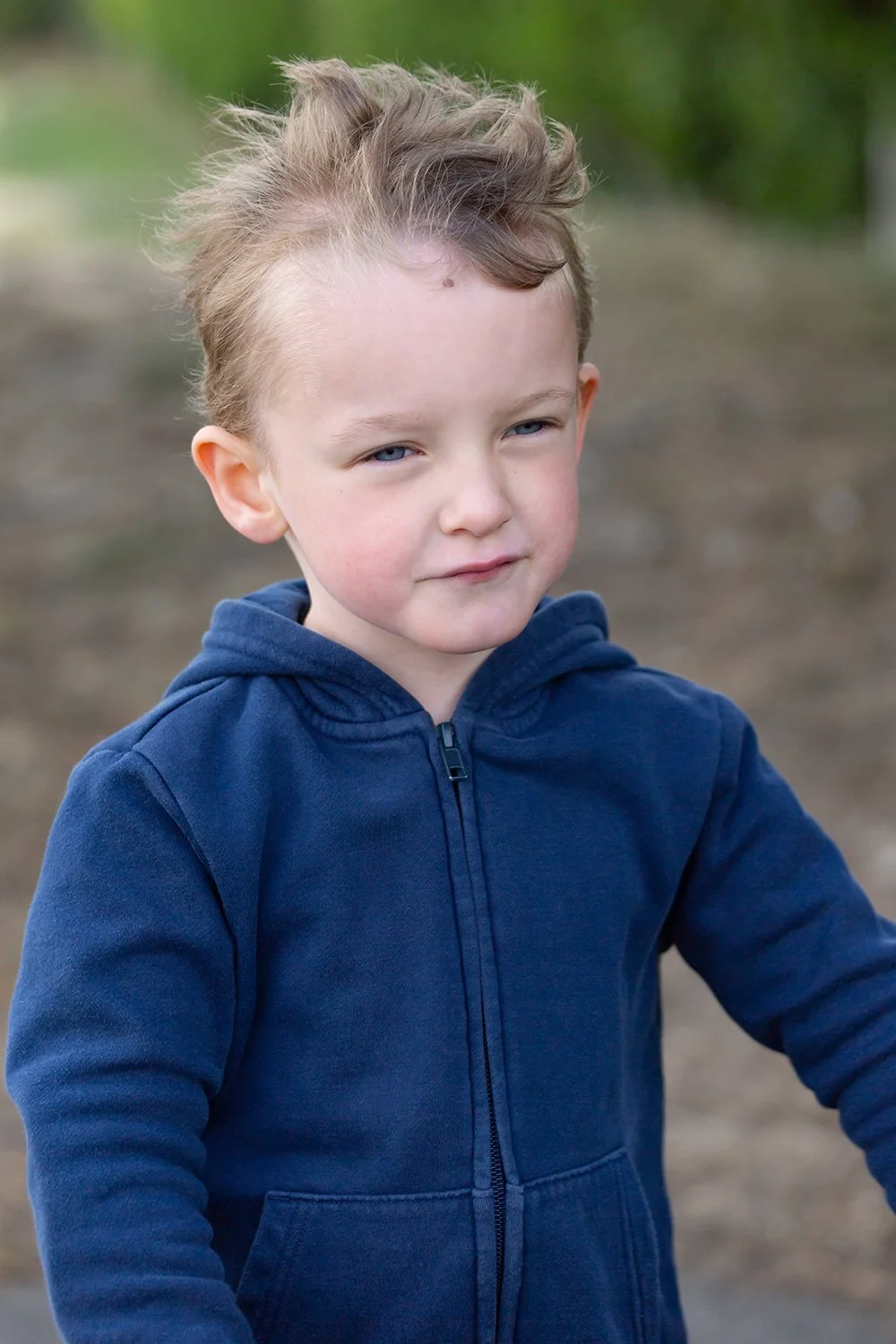 A young boy with tousled blonde hair and blue eyes, wearing a blue zip-up hoodie, outdoors with a blurred natural background.