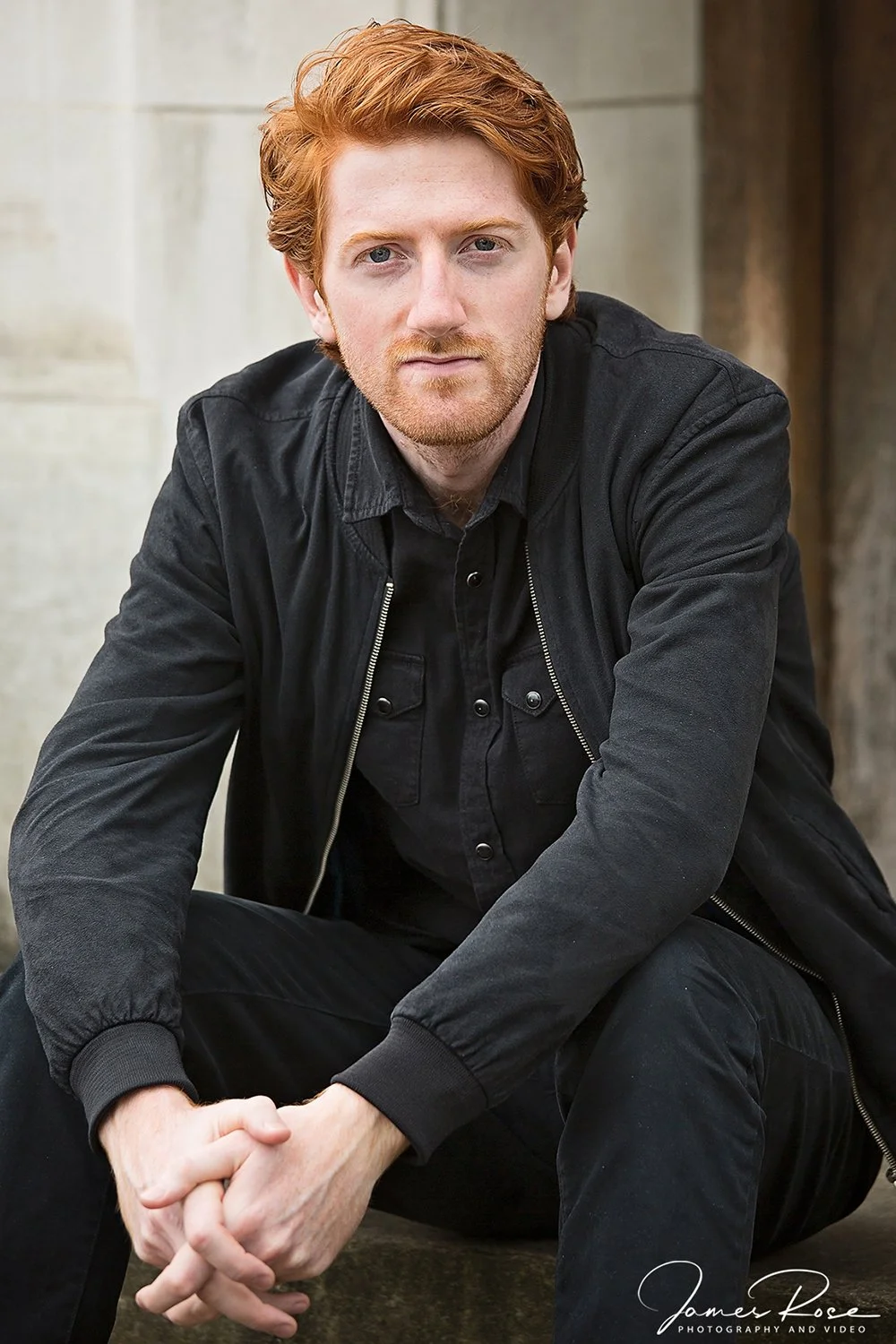 A young man with red hair and beard, wearing a black jacket and black shirt, sitting outdoors against a stone background.