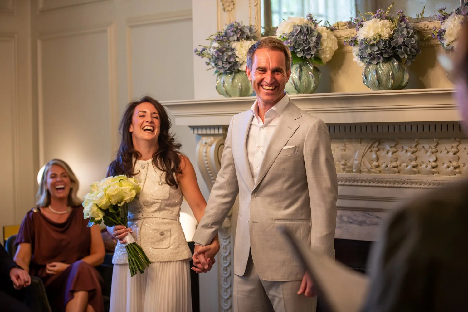 A wedding ceremony with a smiling bride holding a bouquet, standing next to a groom holding her hand, in a decorated room with floral arrangements on the mantle.