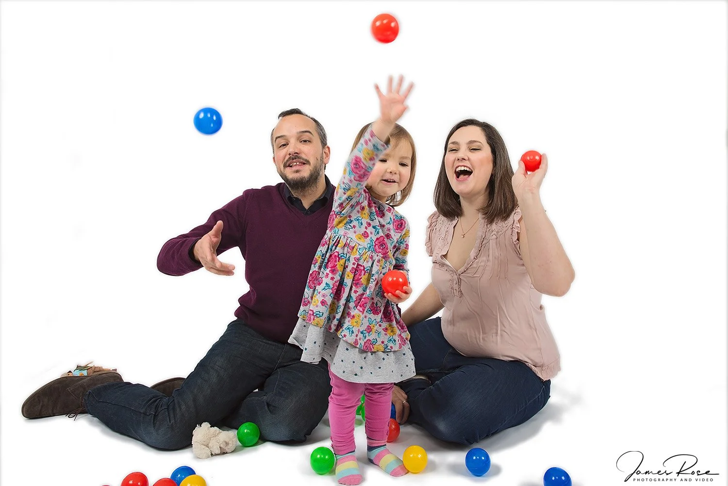 Family of three, two adults and a young girl, playing with colorful plastic balls against a white background. The girl is standing, throwing a red ball, while the adults sit on the floor, smiling and engaging with her.