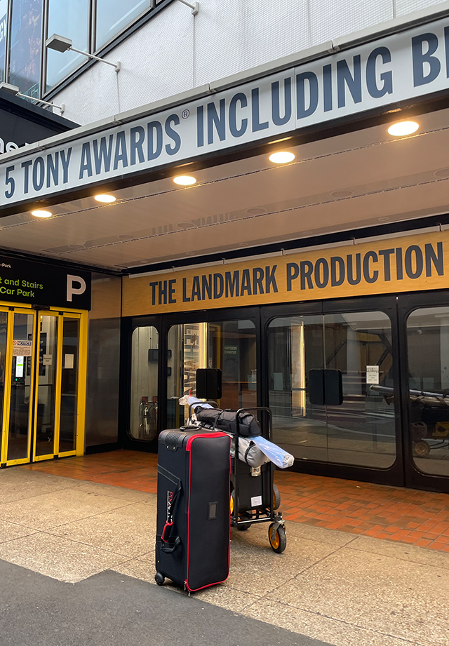 Luggage and a police officer's trolley bag in front of the entrance to a theater called 'The Landmark Production' with signs indicating parking and awards.
