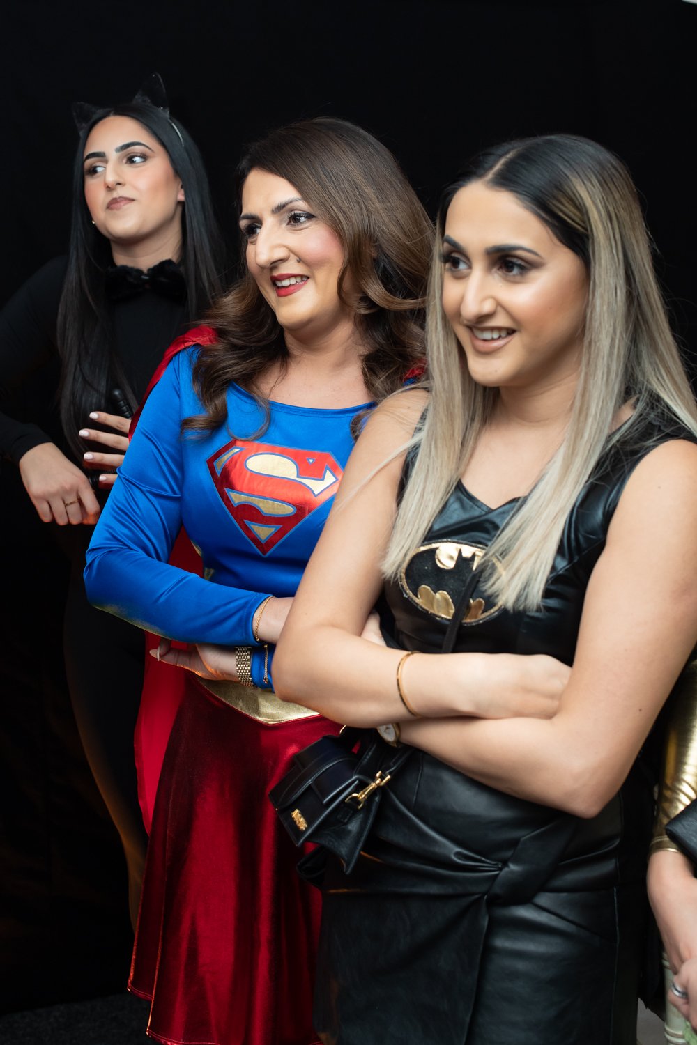 Three women dressed as superhero characters, with one in a Superman costume, one in a Batman costume, and the third woman not in costume, standing together and smiling.