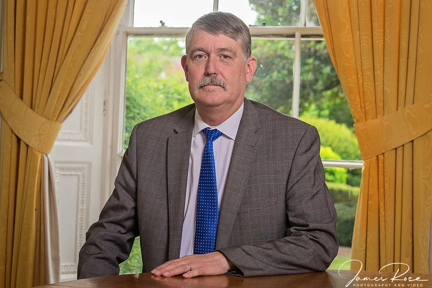 A middle-aged man with gray hair and a mustache, wearing a gray checkered blazer, white shirt, and blue tie, sits at a wooden table in front of a window with yellow curtains, with greenery visible outside.