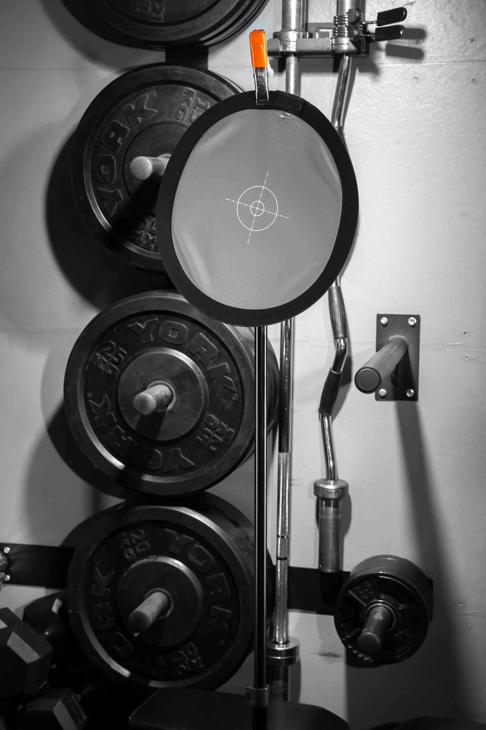 A black and white photo of a workout area with weight plates, a barbell, and a circular target mirror mounted on a stand.