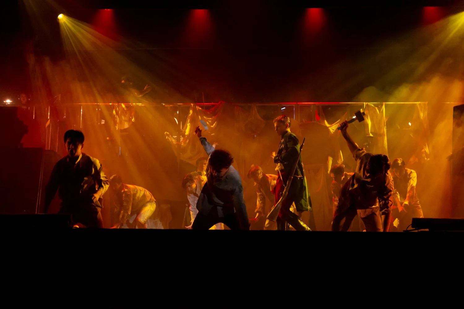 A group of performers on stage with dramatic lighting, dancing and holding props in a theatrical performance.