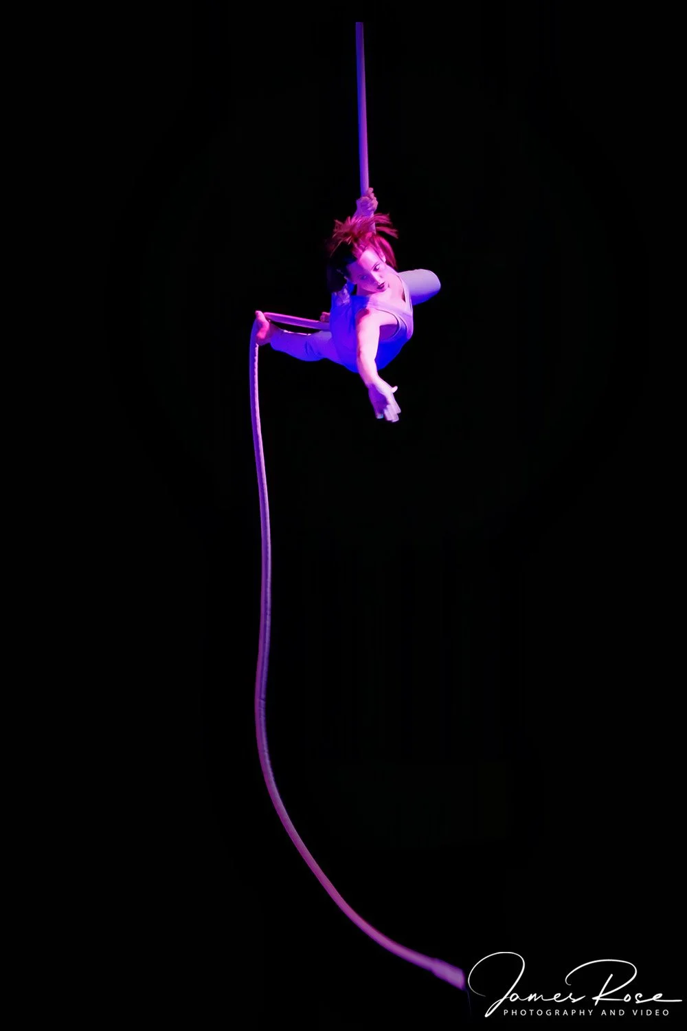 Aerial performer suspended in mid-air with purple lighting, hanging from a long fabric or ribbon, against a black background.