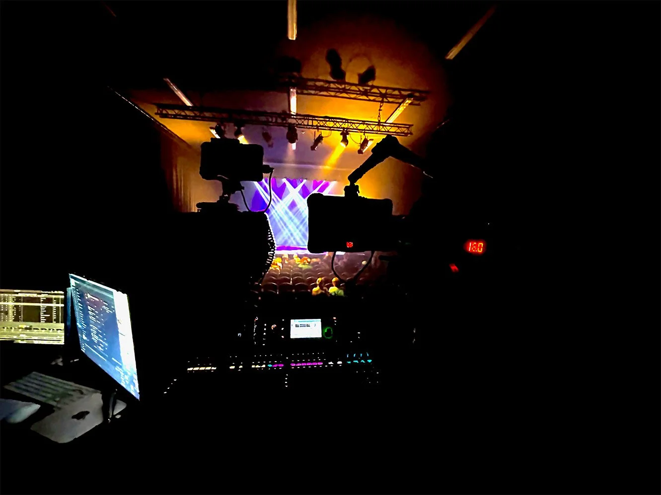 View from behind the sound and lighting control booth overlooking a stage with bright colorful lights, likely at a concert or performance event.